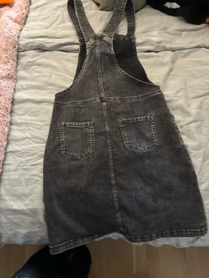 Svart jeansklänning med dragkedja - Cool svart jeansklänning med raka axelband och dragkedja framtill. Klänningen har två stora fickor både fram och bak, och är i en kort modell. Perfekt att styla med en t-shirt eller polotröja under för en avslappnad look.