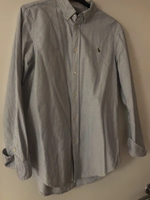 Randig skjorta från Ralph Lauren - Klassisk skjorta från Ralph Lauren med smala blå och vita ränder. Skjortan har lång ärm, knappar framtill och en liten broderad logga på bröstet. Perfekt för en stilren look.