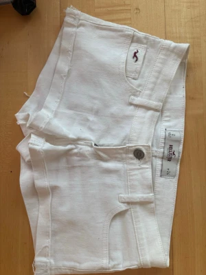 Vita vintage lowwaist 2000s hollister shorts - Snygga vita shorts från Hollister med klassisk femficksdesign och råa kanter. Shortsen har en liten broderad logga i vinrött på fickan och är tillverkade i bomull med en skön passform. Perfekta för varma dagar och enkel att matcha med allt.