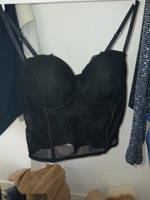 Svart meshkorsett med spetsdetaljer - Snygg svart korsett med meshmaterial och spetsdetaljer över kuporna. Justerbara axelband och markerade sömmar framtill för en extra formande effekt. Perfekt för att skapa en edgy look. Ingen synlig logga eller märke.