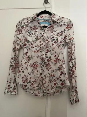Vit blommig skjorta med fickor - Snygg vit skjorta med blommigt mönster i rött, grönt och lila. Skjortan har långa ärmar, två bröstfickor med lock och knappar framtill. 