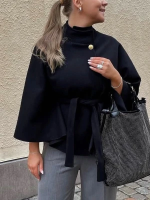 Poncho från missäy  - Stilren svart cape-kappa med hög krage, dekorativ guldknapp och knytskärp i midjan. Vida ärmar och elegant, minimalistisk design. Perfekt för dig som gillar en trendig och sofistikerad look.