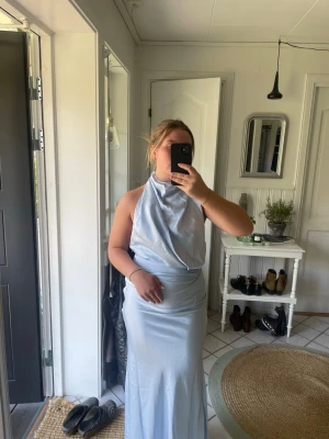 Ljusblå långklänning med halterneck - Snygg ljusblå långklänning med halterneck och draperad överdel. Klänningen har en elegant, slät siluett och är ärmlös med öppen rygg. Perfekt för speciella tillfällen!