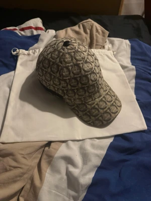 Beige mönstrad keps från Ferregamo  - Ferragamo Interlocking Gancini-keps i beige och svart med det ikoniska monogrammönstret över hela designen. En stilren baseballkeps med böjd skärm och exklusiv premiumkänsla som kombinerar modern streetwear med italiensk lyx. Äkta Kom dm vid frågor den är lite solblekt vid kanten av skärmen inget man märker av där av priset KVITTO FINNS NYPRIS 3500. Dustbag ingår ! 🫡