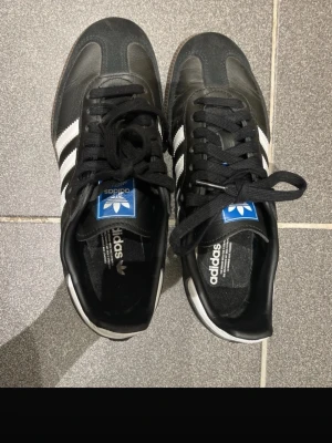 Adidas Samba svartvita sneakers - Klassiska Adidas Samba sneakers i svart läder med vita detaljer och bruna gummisulor. Skorna har de ikoniska tre ränderna på sidorna och blå Adidas-logga på plösen. Perfekt för dig som gillar retrostil och streetwear. Lite slitna längs bak men inget man tänker på