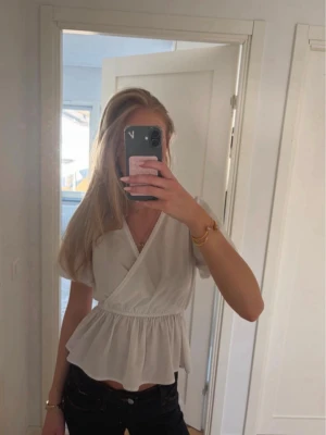 Vit omlottopp med volang - Söt vit omlottopp med korta ärmar och volang nertill. Toppen har v-ringning och resår i midjan som ger en fin passform. Perfekt att styla med jeans eller kjol för en fräsch look.