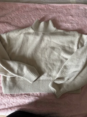 Beige stickad tröja med polokrage - Mysig beige stickad tröja med hög polokrage och breda ribbade muddar vid ärmslut och nederkant. Tröjan har en avslappnad passform och är perfekt för lager-på-lager-stil. Passar dig som gillar enkel och stilren design.