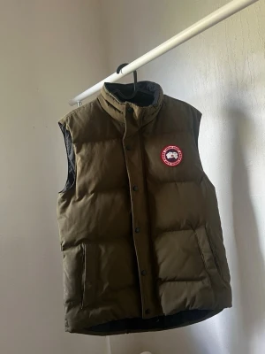 Olivgrön dunväst från Canada Goose - Snygg olivgrön dunväst från Canada Goose med hög krage och tryckknappar framtill. Västen har två stora fickor och den klassiska röda loggan på bröstet. Perfekt för lager-på-lager och håller dig varm under kyliga dagar.