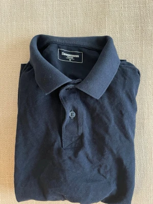 Mörkblå pikétröja från Dressmann - Klassisk mörkblå pikétröja från Dressmann med krage och knappar framtill. Tröjan har korta ärmar och är tillverkad i ett mjukt bomullsmaterial. 