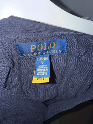 Mörkblå kabelstickad tröja Polo Ralph Lauren - Snygg mörkblå kabelstickad tröja från Polo Ralph Lauren i storlek L (14-16). Tröjan har klassisk rund halsringning och ribbade muddar vid ärmslut och nederkant. Perfekt för dig som gillar tidlös och stilren design.