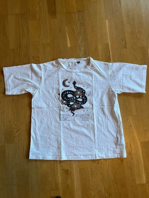 Vit t-shirt med ormmotiv - Vit t-shirt i bomull med coolt tryck av en svart orm, måne och svampar på bröstet. Klassisk rund hals och korta ärmar. Perfekt för dig som gillar unika prints och vill sticka ut lite extra.