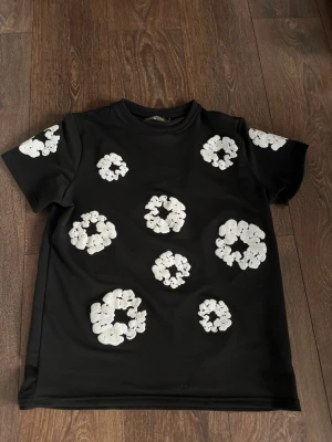 Svart t-shirt med vita blommor - Cool svart t-shirt från Denim tears med vita blommiga mönster över hela tröjan. Klassisk passform med rund hals och korta ärmar. Perfekt för dig som vill sticka ut med en unik och trendig look.