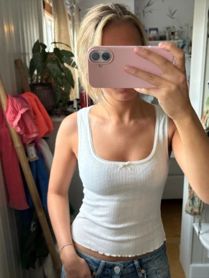 Vit ribbad linnetopp med rosett - Snygg vit ribbad linnetopp med liten rosett framtill och fyrkantig ringning. Toppen är ärmlös och har en croppad passform som sitter tajt mot kroppen. Perfekt att styla med jeans eller kjol för en fräsch look.