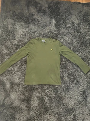 Olivgrön långärmad topp Lyle & Scott - Olivgrön långärmad topp från Lyle & Scott med rund halsringning och klassisk gul logga broderad på bröstet. Tillverkad i mjuk bomull som är skön mot huden. Perfekt att styla med jeans eller kjol för en clean look.