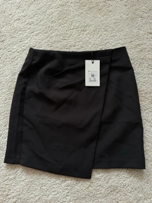 Svart omlottkjol från Bik Bok - Stilren svart kortkjol från Bik Bok med omlottdesign. Kjolen har en enkel och modern look som passar perfekt till många olika outfits. Tillverkad i ett mjukt och bekvämt material som känns skönt mot huden.