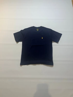 Mörkblå t-shirt från Ralph Lauren storlek M - Snygg mörkblå t-shirt från Ralph Lauren med klassisk gul broderad logga på bröstet. T-shirten har rund halsringning och korta ärmar, perfekt för en stilren och enkel look. Tröjan är helt ny och aldrig buren och har storlek M