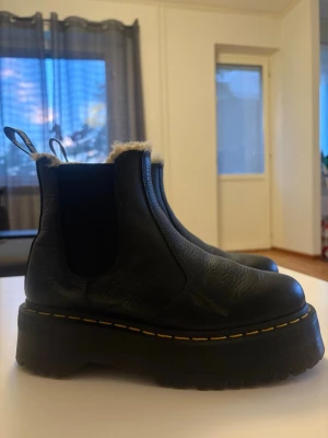 Svarta Dr. Martens Chelsea boots - Snygga svarta Chelsea boots från Dr. Martens med grov platåsula och klassisk gul söm. Skorna är fodrade med mjukt, ljust fuskpälsfoder och har elastiska paneler på sidorna samt dragflikar med logga baktill. Perfekta för kalla dagar!