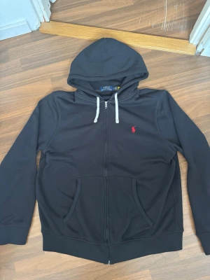 Polo Ralph Lauren - Zip Up Hoodie - Svart hoodie från Polo Ralph Lauren med dragkedja, vit dragsko i huvan och röd broderad logga på bröstet. Kängurufickor framtill och ribbade muddar vid ärmar och nederkant. Tillverkad i mjukt bomullsmaterial för en skön känsla.