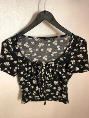 Blommig croppad topp med knyt - Söt svart croppad topp med vita blommor och knyt framtill. Toppen har korta ärmar och en lätt rynkad detalj vid bysten. Perfekt för dig som gillar en feminin och trendig stil.