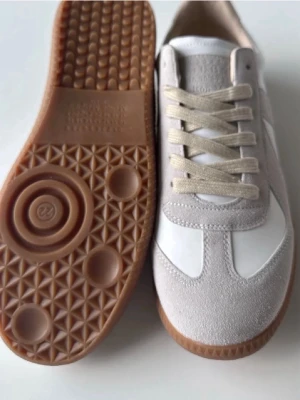 Vita och beige sneakers med gummisula - Stilrena sneakers i vitt och beige med ovandel i läder och mocka, samt klassisk gummisula i brunt. Skorna har beige snörning och en tidlös design som passar till många outfits. Perfekta för dig som gillar minimalistisk och clean stil.