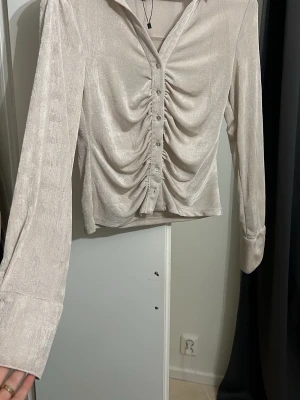 Beige glansig blus med rynk framtill - Trendig beige blus med glansigt tyg och snygg rynkdetalj längs knäppningen framtill. Långa ärmar med breda manschetter och klassisk krage. Perfekt för dig som gillar en stilren men ändå iögonfallande look.