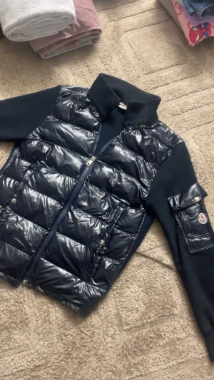 Svart pufferjacka från Moncler - Snygg svart pufferjacka från Moncler med glansig vadderad front och stickade ärmar. Jackan har hög krage, dragkedja framtill och en ficka med Moncler-logga på ena ärmen. Perfekt för kalla vinterdagar och riktigt trendig look.