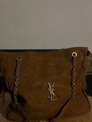 Brun väska från Yves Saint Laurent - Snygg brun handväska från Yves Saint Laurent med guldiga kedjehandtag och YSL-logga framtill. Väskan har en mjuk, fyrkantig form och detaljer i guldmetall. Perfekt för dig som gillar lyxiga accessoarer med trendig känsla.