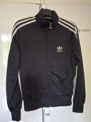 Svart Adidas track jacket - Klassisk svart track jacket från Adidas med silver ränder längs ärmarna och broderad logga på bröstet. Jackan har dragkedja framtill, hög krage och två fickor med dragkedja. Perfekt för sport eller streetwear. Storlek 38