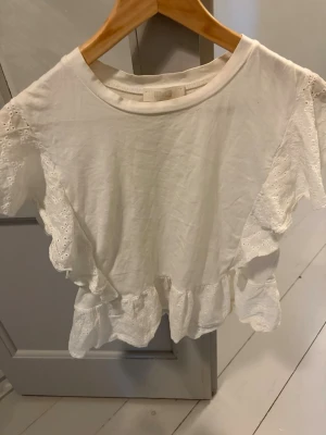 Vit topp med volang och spetsdetaljer - Söt vit topp med volang längs ärmar och nederkant samt fina spetsdetaljer. Toppen har rund halsringning och är kortärmad. Perfekt för dig som gillar romantisk och feminin stil.