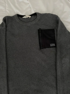 Grå stickad tröja med svart ficka - Säljer en grå stickad tröja från H&M med en svart bröstficka med dragkedja och vit textdetalj. Tröjan har rund halsringning och ett grovt, rutigt stickmönster. Perfekt för dig som gillar en stilren och modern look.