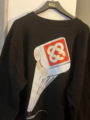 Aldrig använd Svart sweatshirt från Casablanca - svart sweatshirt från Casablanca med stort grafiskt tryck av en pelare och logga i rött, vitt och gult på ryggen. Rund halsringning och avslappnad passform. Perfekt för dig som gillar streetwear och unika prints.