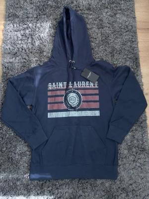 Mörkblå hoodie från Saint Laurent - Snygg mörkblå hoodie från Saint Laurent med tryckt logga och ränder framtill. Klassisk känguruficka och justerbar huva med snören. Perfekt för en avslappnad och trendig stil. Materialet känns mjukt och bekvämt mot huden.