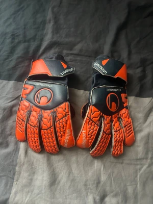 Uhlsport orangea målvaktshandskar - Säljer ett par grymma målvaktshandskar från Uhlsport i orange och svart. Handskarna har coola detaljer och justerbart kardborreband vid handleden för bästa passform. Perfekta för dig som älskar fotboll och vill ha stil på planen.