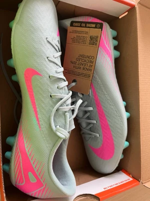 Nike fotbollsskor ljusgrå/rosa - Snygga fotbollsskor från Nike i ljusgrått med neonrosa detaljer och logga. Skorna har snörning, mönstrad ovandel i syntetmaterial och turkosa dobbar för bra grepp på planen. Perfekta för dig som vill sticka ut på fotbollsplanen. Oanvända med kvitto från XXL