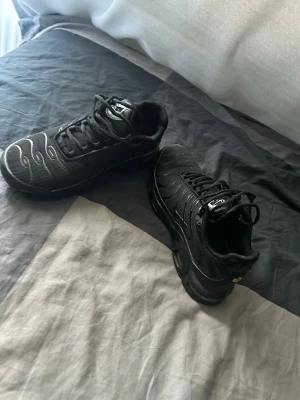 Svarta Nike Air Max Plus sneakers - Säljer ett par svarta Nike Air Max Plus sneakers med klassiska vågiga detaljer på ovandelen och synlig Air Max-dämpning i sulan. Skorna har mesh och syntetmaterial, svarta skosnören och diskreta loggor. Perfekta för dig som gillar streetwear och sportig stil.