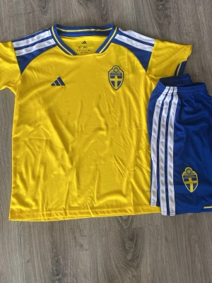 Sverige fotbollströja och shorts Adidas - Gult och blått matchställ från Adidas med Sveriges landslagslogga på både tröja och shorts. Tröjan har korta ärmar, blå detaljer och vita ränder på axlarna. Shortsen är blå med vita ränder och resår i midjan. Perfekt för fotbollsträning eller match.