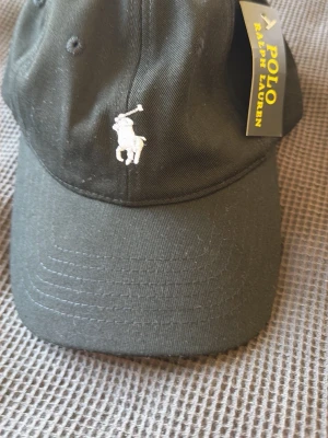 Svart keps från Polo Ralph Lauren - Svart keps från Polo Ralph Lauren med klassisk böjd skärm och broderad vit logga med polospelare framtill. Justerbar rem baktill med metallspänne där det står 'POLO'. Perfekt accessoar för en sportig och stilren look.