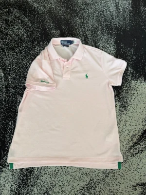Ljusrosa pikétröja från Polo Ralph Lauren - Ljusrosa pikétröja från Polo Ralph Lauren med klassisk krage, korta ärmar och grönt broderat logotyp på bröstet samt 'Polo' broderat på ärmen. Perfekt för en stilren och sportig look.