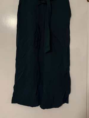 Mörkgröna vida byxor med knytband - Snygga mörkgröna byxor med hög midja och knytband i midjan. Byxorna har en rak och vid passform som ger en avslappnad look. Perfekta för dig som gillar stilrena och bekväma plagg. Materialet känns mjukt och lätt.