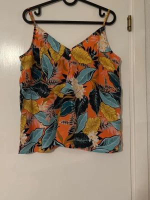 Blommig linnetopp från Gina Tricot - Färgstark linnetopp från Gina Tricot med tropiskt blommönster i orange, gul, blå och svart. Toppen har smala axelband, v-ringning och dekorativa knappar framtill. Perfekt för varma dagar och ger en riktigt somrig vibe.