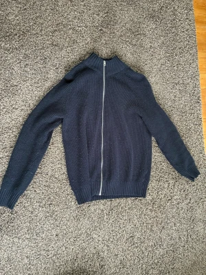 Mörkblå stickad ziptröja - Mörkblå stickad tröja med dragkedja framtill och hög krage. Tröjan har ribbade muddar vid ärmslut och nederkant, samt en klassisk rak passform. Perfekt för lager-på-lager och enkel att matcha med jeans eller chinos.