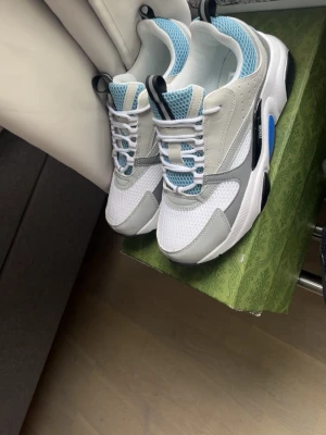 Puma RS-X sneakers vit/blå/grå - Snygga Puma RS-X sneakers i vitt, grått och blått med svarta detaljer. Ovandel i mesh och syntet, chunky sula och cool design. Skorna har blå meshpaneler, vita snören och svart logga på sidan. Perfekta för en sportig och trendig look.