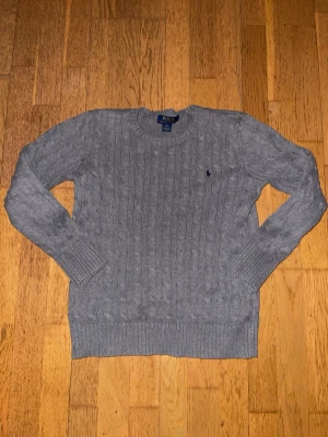 Grå kabelstickad tröja Polo Ralph Lauren - Kabelstickad grå tröja från Polo Ralph Lauren med rund halsringning och diskret broderad logga på bröstet. Långa ärmar och ribbade muddar ger en klassisk look. Perfekt för lager-på-lager och enkel att matcha med jeans. Tröjan passar en som är 165 cm  lång ungefär. 