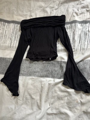 Svart offshoulder topp från SHEIN - Trendig svart croppad, offshoulder topp från SHEIN med långa utsvängda ärmar. Toppens material är mjukt och stretchigt, perfekt för en cool och avslappnad stil. Passformen är tight och toppen har en croppad längd som ger en modern vibe.