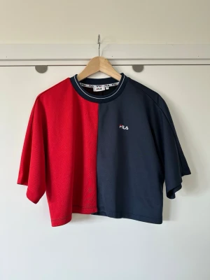 Röd och marinblå croppad FILA t-shirt, storlek M - Cool FILA T-shirt i storlek M med en design som är uppdelad i rött och mörkblått. Tidlös modell med rund halsringning med en vit randdetalj och en liten Fila-logotyp broderad på bröstet. Baksidan av t-shirten har en stor Fila-logotyp i vitt och mörkblått. Ärmarna är korta och vida, vilket ger en avslappnad passform. Finvävt funktionstyg / sporttyg, så bra material som andas. Mycket fint skick, som ny!