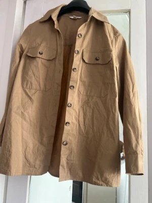 Beige overshirt med fickor - Stilren beige overshirt med klassisk krage och knäppning framtill. Två bröstfickor med lock och knapp ger en cool utility-look. Långärmad och tillverkad i ett lätt bomullstyg, perfekt för lager-på-lager-stil.