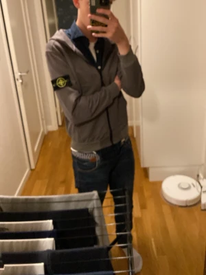 Grå/grön hoodie från Stone Island - Säljer en grå/grön hoodie från Stone Island med dragkedja framtill och klassisk logotyp-patch på vänster ärm. Hoodien har huva och ribbade muddar vid ärmslut och nederkant. Perfekt för en avslappnad och stilren look.