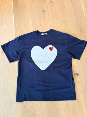 Blå t-shirt från Comme des Garçons - Mörkblå t-shirt i bomull från Comme des Garçons Play med stort vitt hjärta och rött hjärta med ögon på bröstet. Klassisk passform och rund halsringning. Perfekt för dig som gillar streetwear och unika prints.