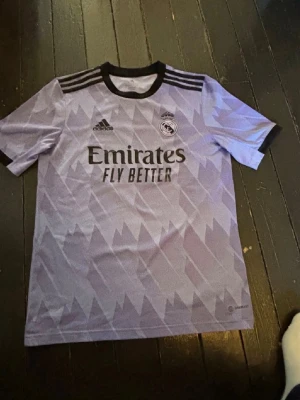 Lila Real Madrid matchtröja Adidas - Säljer en lila Real Madrid matchtröja från Adidas med svart krage och svarta detaljer. Tröjan har ett grafiskt mönster, klubbmärke och Adidas-logga på bröstet samt Emirates Fly Better-tryck. Tillverkad i lätt och ventilerande material, perfekt för fotbollsträning.