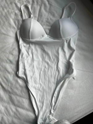 Vit body med bygel och vadderade kupor - Säljer en stilren vit body med justerbara axelband, bygel och vadderade kupor för extra stöd. Bodyn har en slät och stretchig passform med knäppning i grenen. Perfekt för dig som gillar en enkel och klassisk look. Materialet känns mjukt och följsamt mot huden. Oanvänd! 